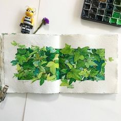 an open book with green leaves on it next to a small toy and watercolor paints