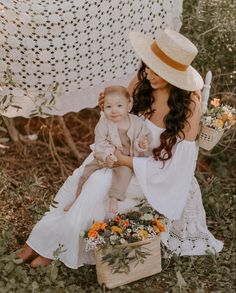 a woman in a white dress and hat holding a baby next to a basket with flowers