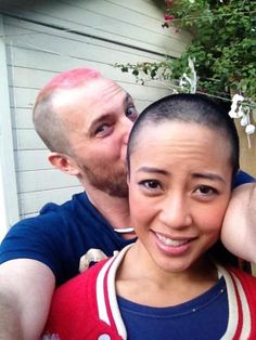 a man and woman taking a selfie in front of a house with pink hair