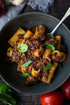 a bowl filled with pasta and meat on top of a wooden table next to tomatoes