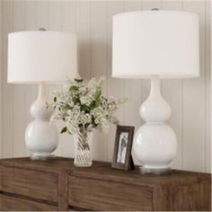 two white vases with flowers sit on a dresser next to a framed photograph and lamp