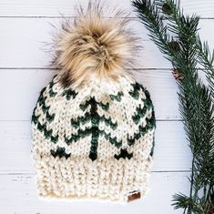 a white and green knitted hat next to pine branches on a white wooden surface