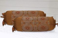 two decorative pillows sitting on top of a white bed covered in brown and red fabric