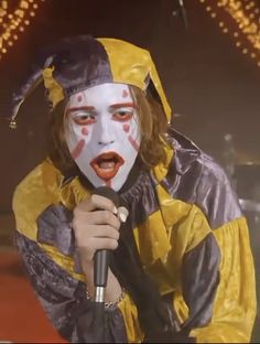 a man in clown makeup holding a microphone and wearing a yellow outfit with red paint on his face