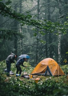 two people standing next to a tent in the woods with an umbrella over their head