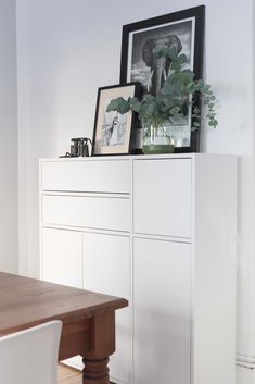 a white cabinet sitting next to a wooden table with pictures on it and a plant in the corner