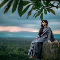 a woman sitting on top of a stone wall next to a lush green forest under a cloudy sky