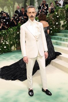 a man in a white tuxedo and black shoes stands on the green carpet