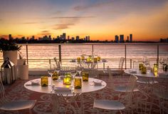 an outdoor dining area overlooking the water and cityscape at sunset, with white chairs and tables set up for dinner