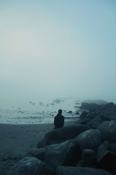 a person sitting on rocks near the ocean with fog in the air and water behind them