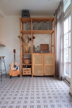 a living room with a guitar on the floor next to a book shelf and window