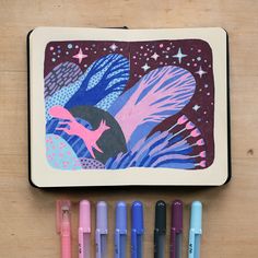 some markers and pens are sitting next to a notebook with an image of a mermaid on it