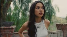 a woman with long black hair standing in front of a tree and brick wall, wearing a white top