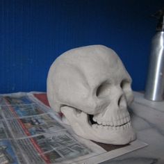 a fake human skull sitting on top of a table next to a cup and newspaper