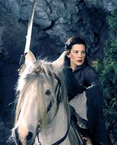 a woman riding on the back of a white horse next to a stone wall and trees