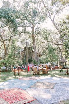 a group of people sitting at tables in the middle of a park with trees and rugs on the ground