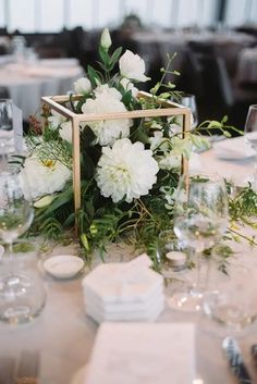 a table with white flowers and greenery in a square vase on top of it