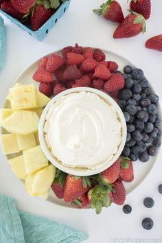 a plate filled with fruit and dip surrounded by strawberries, pineapples, blueberries, and bananas