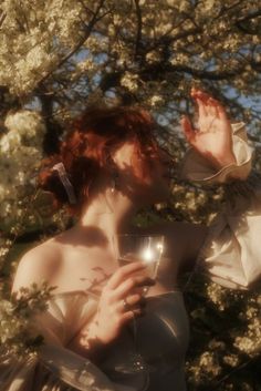 a woman with red hair holding a wine glass in front of her face while standing next to a tree