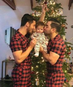 two men are holding a baby in front of a christmas tree