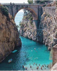 people are swimming in the blue water under an old stone bridge that is connected to a cliff