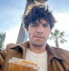 a man holding a drink in his right hand and looking at the camera with palm trees behind him