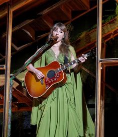 a woman in a green dress holding a guitar and singing into a microphone while standing on stage