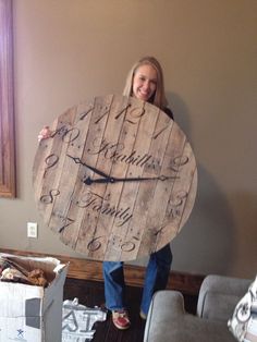 a woman standing next to a wooden clock with writing on the face and numbers painted on it