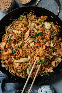 a wok filled with noodles and chicken on top of a table next to two wooden chopsticks