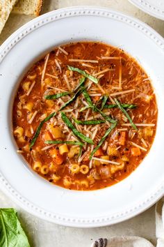 a white bowl filled with pasta soup and garnished with green onions, parmesan cheese and basil