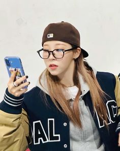 a girl wearing glasses and a baseball cap is looking at her cell phone while standing in front of a white wall