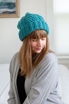 a woman sitting on a bed wearing a blue knitted beanie and looking down at her cell phone