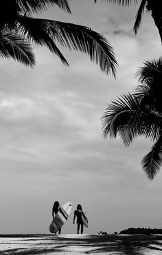 two people carrying surfboards on the beach under palm trees