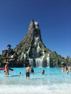 people are swimming in the water near a waterfall at an amusement park, with a mountain behind them