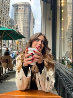 a woman sitting at a table with a cup of coffee in her hand and looking into the camera