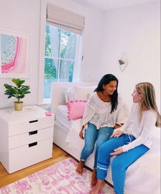 two women sitting on a bed in a room with white walls and pink rugs