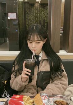 a woman sitting at a table with food and drink in her hand while holding a straw up to her mouth