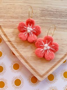 two small pink flowers are hanging from gold earwires on a wooden surface with orange and white circles