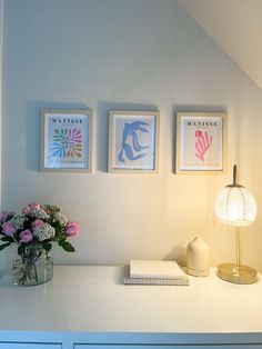 three framed pictures hang on the wall above a white desk with flowers and a lamp