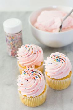 three cupcakes with pink frosting and sprinkles next to a bowl of powder