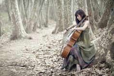 a woman in a long dress holding a violin while standing next to a tree trunk