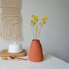 small yellow flowers in a clay vase next to a wooden spoon on a white tablecloth