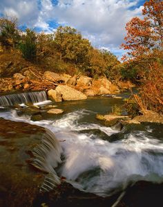 a river running through a forest filled with lots of rocks and trees under a cloudy blue sky