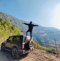 a man standing on the back of a black jeep in front of a mountain range