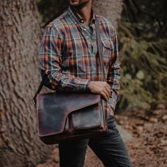 a man in plaid shirt carrying a brown leather messenger bag on forest path with trees