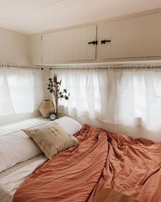 a bed with an orange comforter and pillows in front of two white curtains on the wall