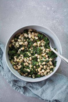 a bowl filled with beans and greens on top of a blue cloth next to a spoon