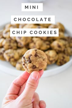 a hand holding up a chocolate chip cookie in front of a white bowl full of cookies