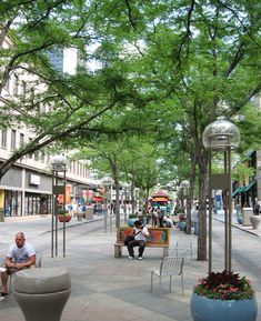 people are sitting on benches in the middle of an open area with trees and buildings