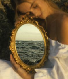 a woman holding a mirror in front of her face with the ocean and rocks behind her